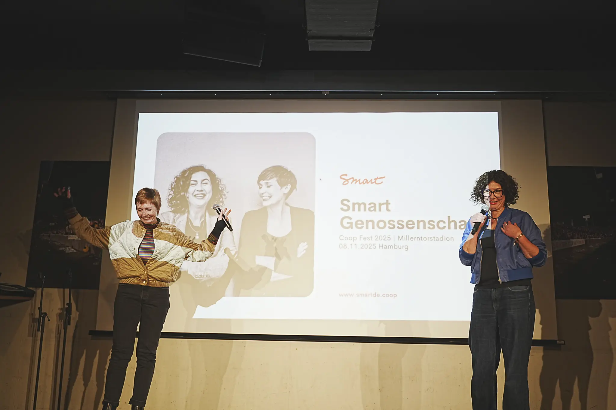 Zwei Frauen auf der Bühne. Im Hintergrund eine Präsentation, auf der Smart Genossenschaft steht.